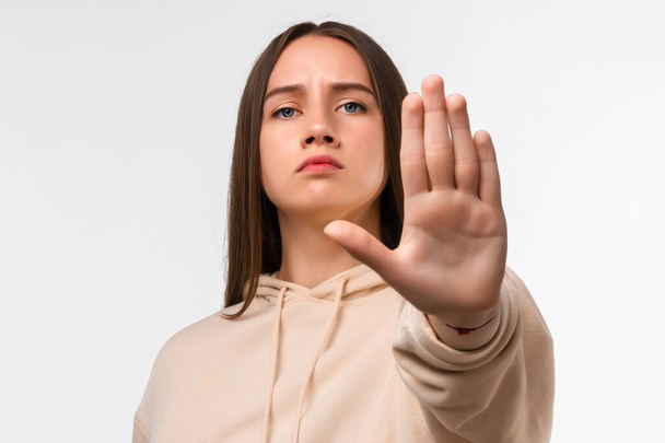 Young pretty woman with long chestnut hair, looking serious and stern, wearing casual beige hoodie, showing open palm making stop gesture - Photo, Image