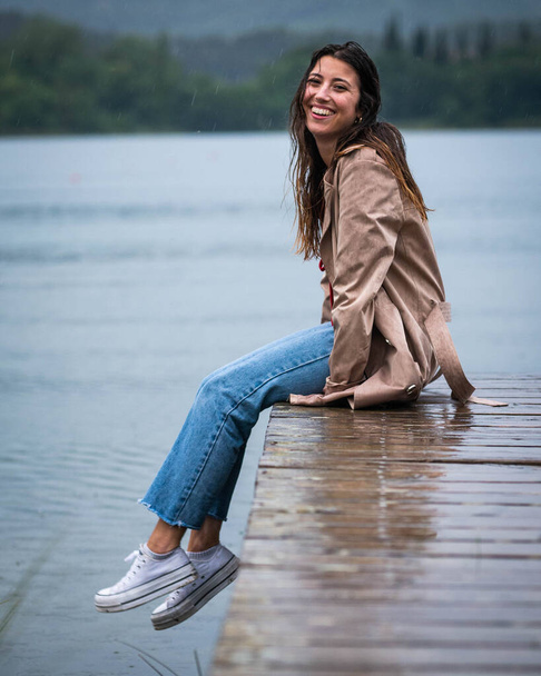 young woman sitting on a wooden pier in the rain in a beautiful lake - Фото, зображення
