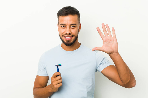 Joven hombre sudasiático sosteniendo una hoja de afeitar sonriente alegre mostrando el número cinco con los dedos. - Foto, Imagen