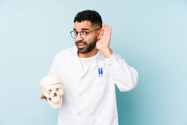 Young doctor latin man holding a skull isolated trying to listening a gossip. - Photo, Image