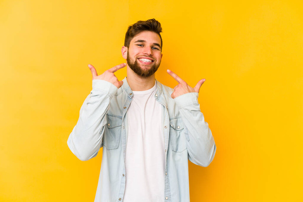 Young caucasian man isolated on yellow background smiles, pointing fingers at mouth. - Photo, Image