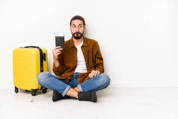 Joven hombre caucásico sentado con un pasaporte y una maleta aislada confundida, se siente dudoso e inseguro. - Foto, Imagen