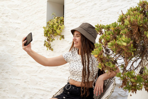 Chica joven tomando una selfie. Alrededor hay vegetación y una pared blanca en el fondo. - Foto, Imagen