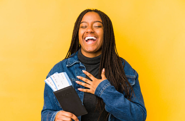 Joven afroamericana americana sosteniendo un pasaporte aislado se ríe en voz alta manteniendo la mano en el pecho. - Foto, imagen