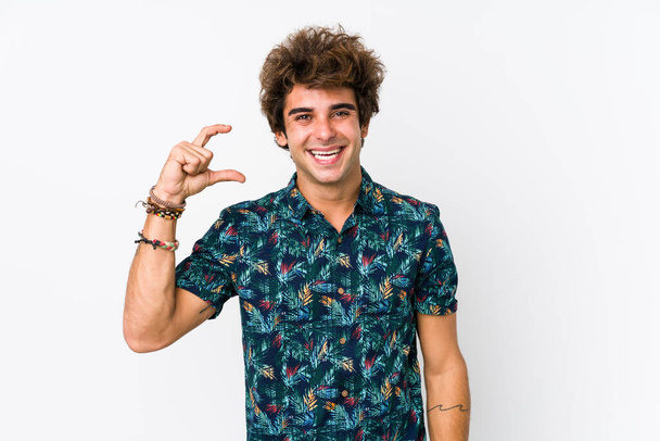Молодой кавказский мужчина в цветочной футболке, изолированно держащий что-то маленькое указательными пальцами, улыбающийся и уверенный. - Фото, изображение