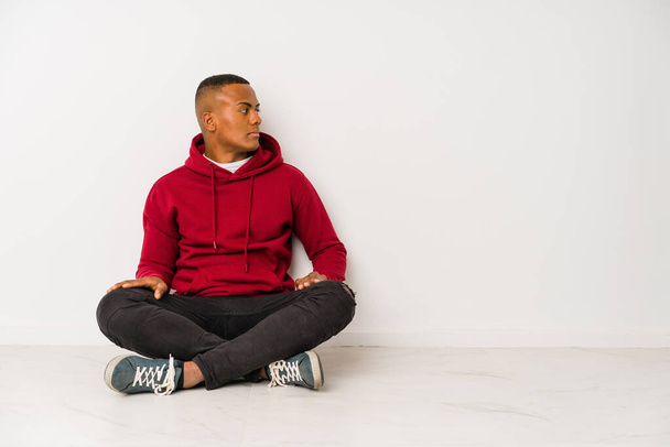 Jovem latino sentado no chão isolado olhando para a esquerda, de lado pose. - Foto, Imagem