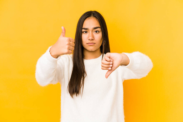 Jonge vrouw geïsoleerd op een gele achtergrond tonen duimen omhoog en duimen omlaag, moeilijk kiezen concept - Foto, afbeelding