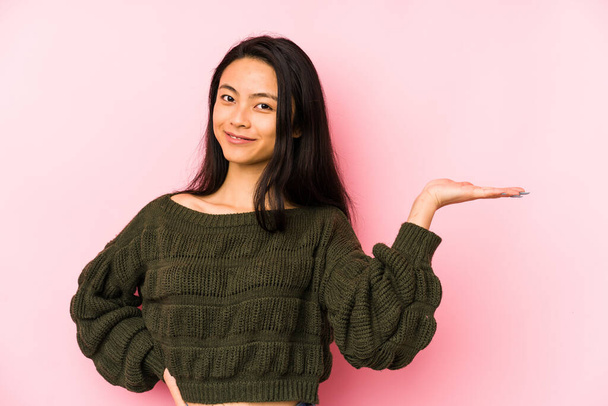 Young chinese woman isolated on a pink background showing a copy space on a palm and holding another hand on waist. - Photo, Image