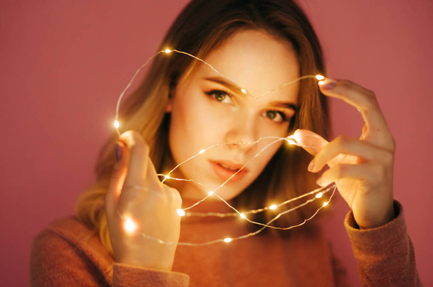 Background. Focus on Christmas lights in the hands of a girl. Photo of a woman with Christmas lights in her hands on a pink background. - Foto, Imagem