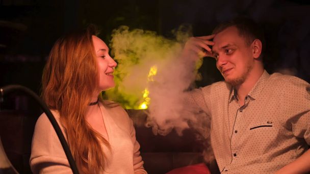 Schönes Paar, das Wasserpfeife raucht und miteinander spricht. Medien. Freund produziert Rauch aus seinem Mund, während er mit seiner Freundin bei einem Date plaudert. - Foto, Bild