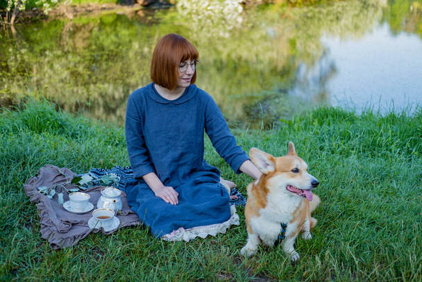 Young woman in retro dress with funny corgi dog on the picnic, female with cute dog drink english tea in the park - Foto, imagen