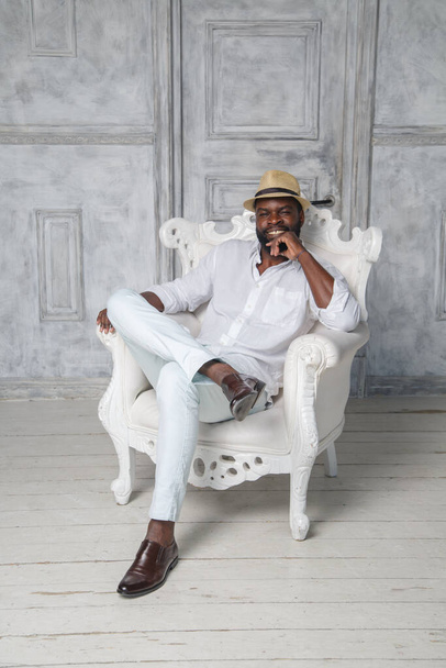 A smiling bearded black elegant man in a white suit and hat sits in a chair in a bright room. - Fotó, kép
