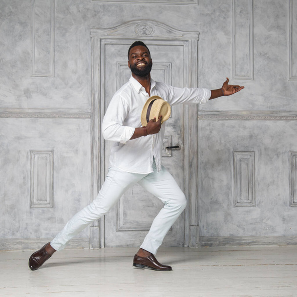 Black elegant man in a white suit is dancing in a bright room near the door holding a hat in one hand. - Foto, Imagen