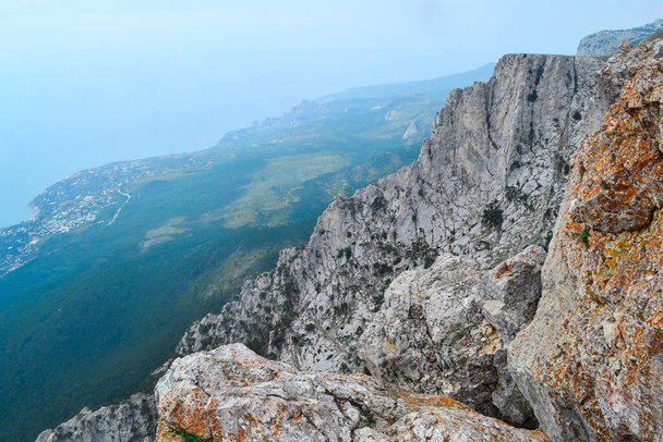 large steep sharp stone rock cliff with red moss against the background of Black sea coastline, the city and forest below, summer - Foto, afbeelding