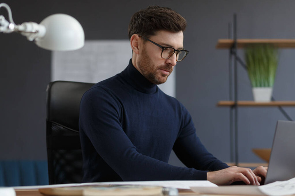Architect working in office with laptop. Engineer thinks over architectural plan, searching new ideas for construction project. Portrait of handsome bearded man sitting at workplace. Business concept - Fotoğraf, Görsel