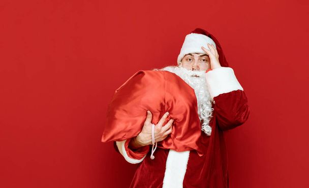 Grappige Kerstman met een tas draagt geschenken voor Kerstmis, studio portret.Portret van een vermoeide Kerstman met een zak in zijn handen op een rode achtergrond, kijkt in de camera en past zijn hoed op zijn hoofd - Foto, afbeelding