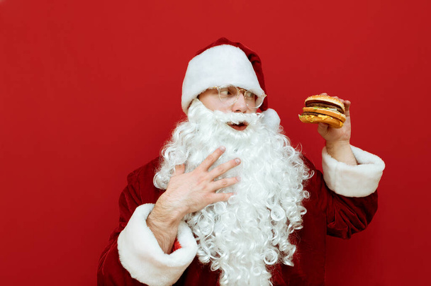 Portrait du Père Noël affamé regardant un hamburger à la main et caressant la barbe isolée fond rouge.Homme drôle en costume de Père Noël debout sur fond rouge avec de la restauration rapide et aller manger. - Photo, image