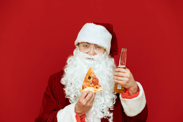 Portrait rapproché d'un homme positif en costume de Père Noël isolé sur fond rouge, tenant une pizza en tranches et une bouteille de bière, regardant dans la caméra et souriant.Le Père Noël mange de la restauration rapide et boit de l'alcool - Photo, image