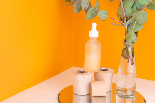 Retinol serum bottle on wooden pedestal and glass vase with eucalyptus branch on orange background. Organic healing cosmetics. Eco skin care. Beauty hyaluronic acid treatment - Fotografie, Obrázek