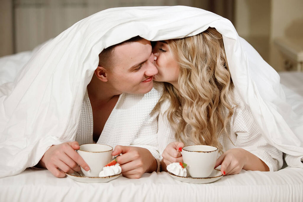 Couple under the covers with a cup of coffee in bed. Lovers smile, enjoy the tranquility. Romantic idyll, love relationships. - Photo, Image