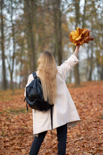 Стильная девушка в модном осеннем пальто, черных маминых джинсах и кожаном черном рюкзаке с букетом осенних листьев в осеннем парке. Вид со спины.                                  - Фото, изображение
