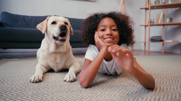 Afrikaans amerikaans meisje zwaaien hand in de buurt hond liggen op tapijt thuis - Video