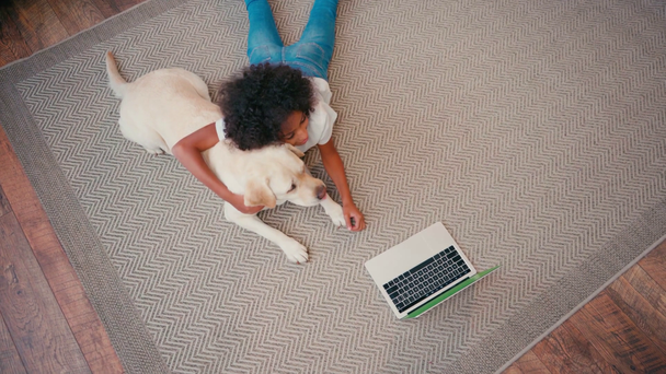 Overhead view of african american girl hugging dog while lying on carpet at home - Footage, Video