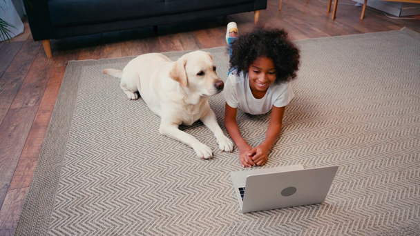 African American κορίτσι που βρίσκεται κοντά στο σκυλί και κοιτάζοντας φορητό υπολογιστή στο χαλί στο σπίτι - Πλάνα, βίντεο