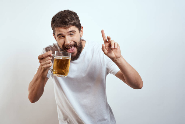 man with a mug of beer in his hands and a white t-shirt light background mustache beard emotions model - Foto, imagen