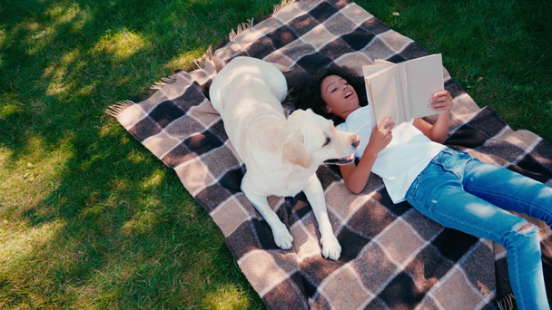 Afričanky americká dívka čtení knihy, zatímco leží na kostkované přikrývce poblíž labrador  - Záběry, video