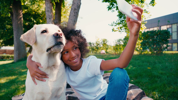 Afrikkalainen amerikkalainen tyttö halaus koira, ottaen selfie ja istuu ruudullinen huopa - Materiaali, video
