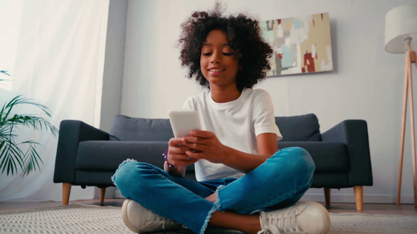 Chica afroamericana con las piernas cruzadas usando el teléfono celular mientras está sentado en la alfombra - Imágenes, Vídeo