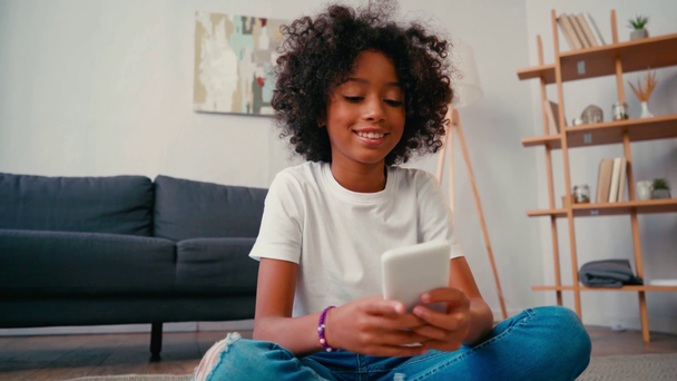 Африканская американская девушка сидит на полу и пишет смс на мобильный телефон дома - Кадры, видео