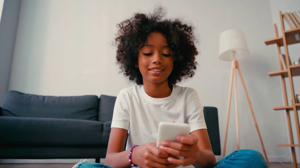 Африканская американка пишет смс на смартфоне, сидя дома на полу - Кадры, видео