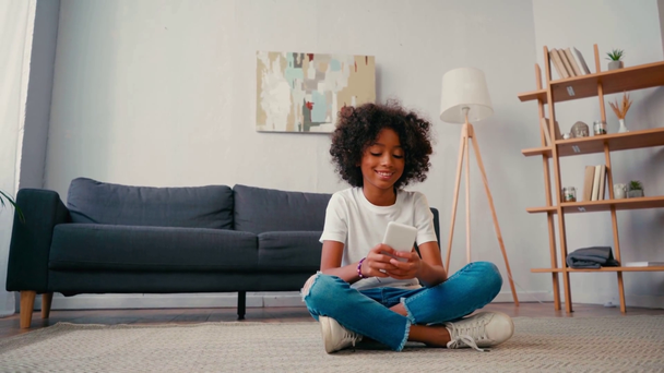 Afrikai amerikai lány keresztbe tett lábakkal sms-ezik mobilon otthon - Felvétel, videó