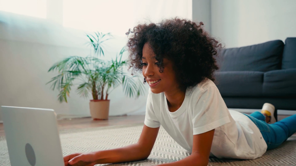 Africano menina americana deitada no tapete perto do sofá e usando laptop em casa - Filmagem, Vídeo