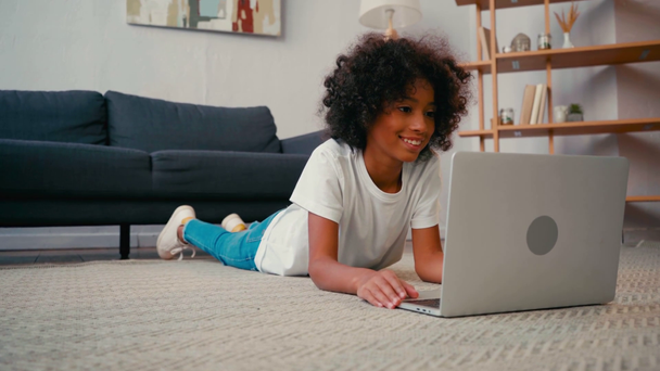 Smiling african american girl lying on carpet and looking at laptop at home - Footage, Video