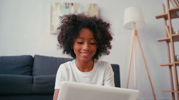Hymyilevä afrikkalainen amerikkalainen tyttö käyttää digitaalista tablettia istuessaan lähellä sohvaa - Materiaali, video