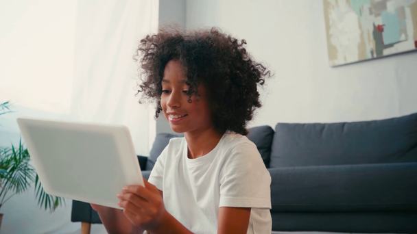 African american girl with crossed legs using digital while sitting near couch - Séquence, vidéo