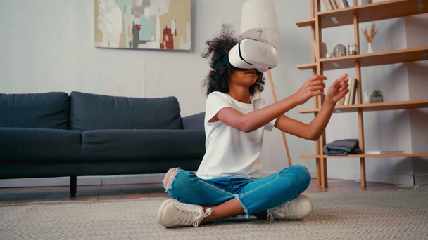 Африканская девочка со скрещенными ногами жестикулирует, используя VR-гарнитуру дома - Кадры, видео