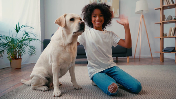Afrikaans amerikaans meisje strelen hond en zwaaien op camera terwijl zitten op tapijt - Video