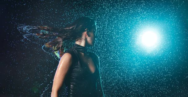 Beautiful wet girl in a swimsuit shakes her head and streams of water flow down from her hair under raindrops with turquoise highlight on a black background. - 写真・画像
