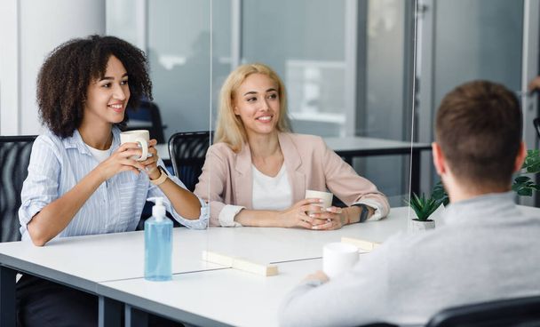 Smiling european and african american businesswomen with cups communicating with guy through glass partition - Photo, Image