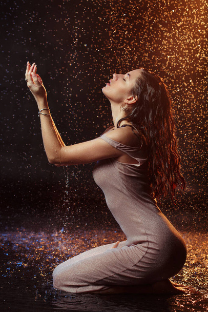 Beautiful sexy girl with drops of water on her face in splashes illuminated by an orange light against a dark background in the studio. - Φωτογραφία, εικόνα