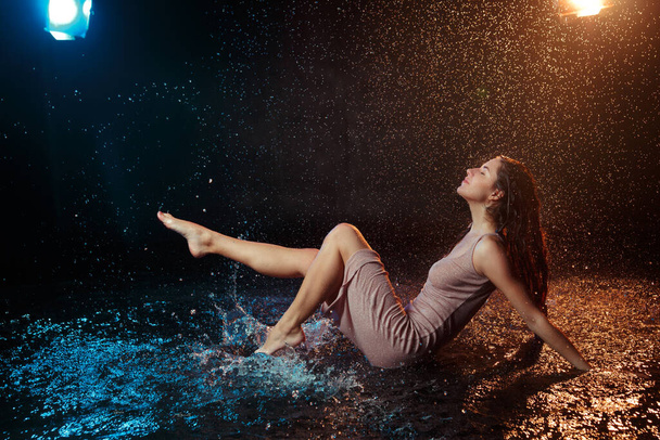 Beautiful sexy girl with drops of water on her face in splashes illuminated by an orange light against a dark background in the studio. - Фото, зображення