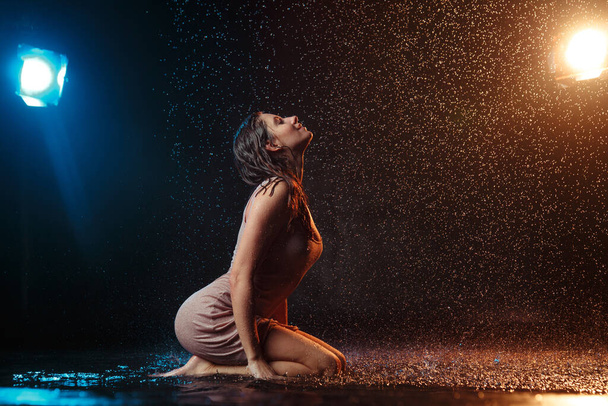 Beautiful sexy girl with drops of water on her face in splashes illuminated by an orange light against a dark background in the studio. - Photo, image