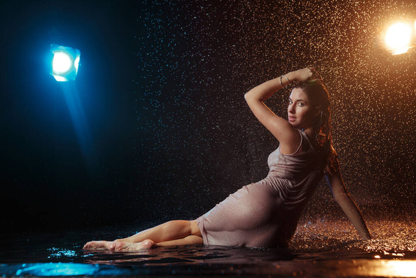 Beautiful sexy girl with drops of water on her face in splashes illuminated by an orange light against a dark background in the studio. - Foto, Imagen
