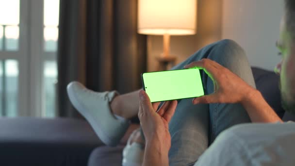 Evdeki adam kanepede uzanıyor ve yatay modda yeşil ekran ile akıllı telefon kullanıyor. İnternete bakıyor, içeriği, videoları, blogları izliyor. - Video, Çekim