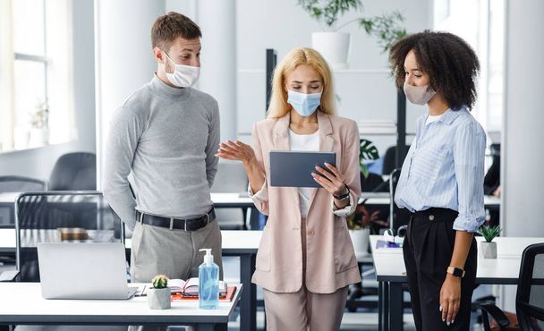 Questions to manager during work. Woman boss in protective mask looks at tablet and checks task from guy and african american lady - Photo, Image