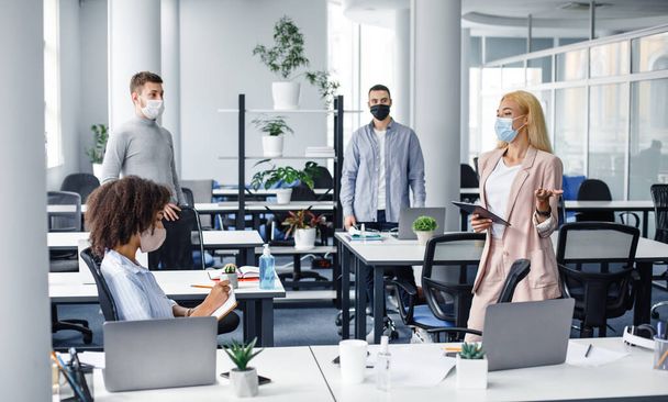 Встреча в офисе и инструкции от босса. Молодая деловая женщина в защитной маске с планшетом в руке разговаривает с работниками на рабочем месте - Фото, изображение